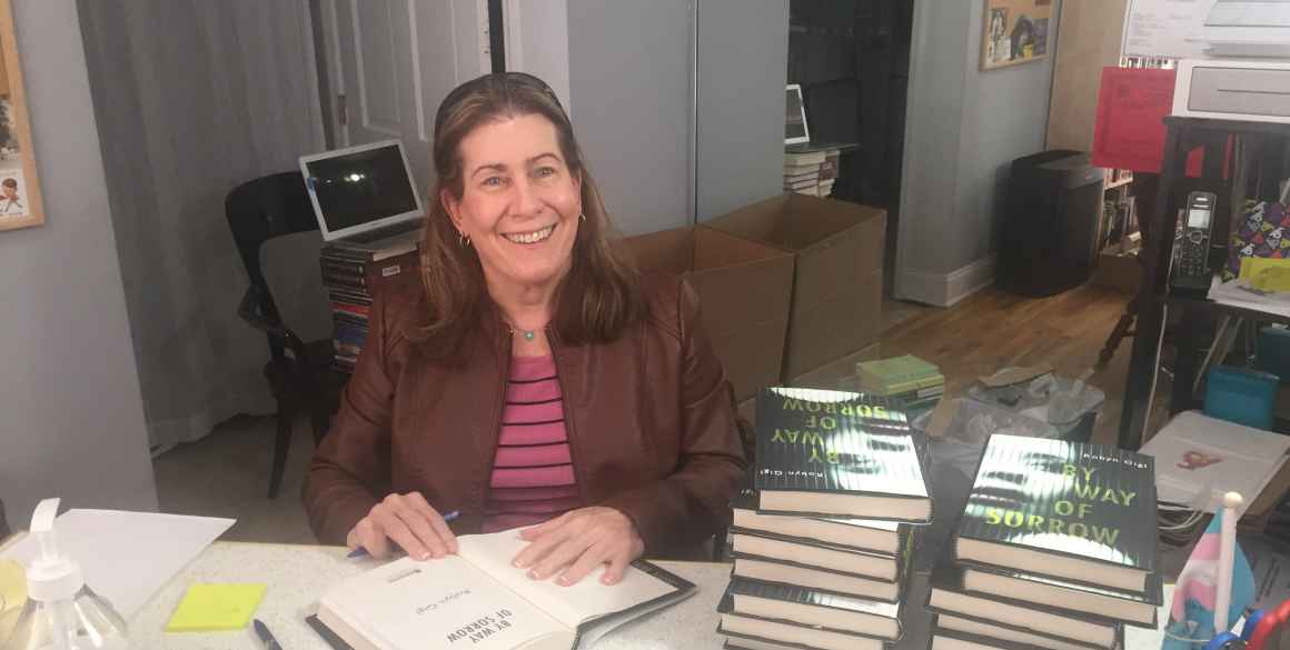 Robyn Gigl smiles for the camera as she sits behind a table of stacked books to her left, and has a book open to sign it.