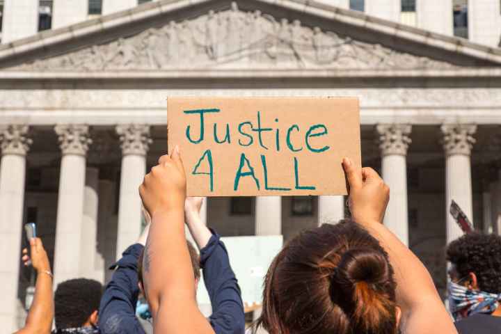 Protester holding sign reading "Justice 4 ALL" in front of state building