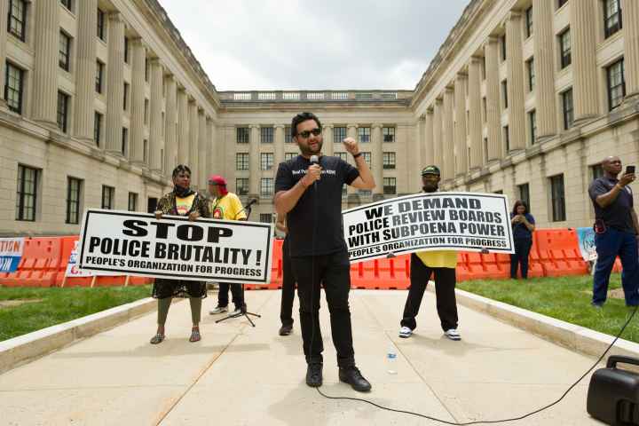 Amol SInha is speaking into a microphone at the State House. Two people are standing behind him holding signs that read "stop police brutality" and "we demand  police review boards with subpoena power"