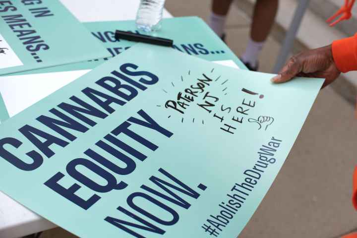 A mint green sign reads: Cannabis equity now. Hashtag Abolish the Drug War. A handwritten note is written off the side that reads: Paterson NJ is here!