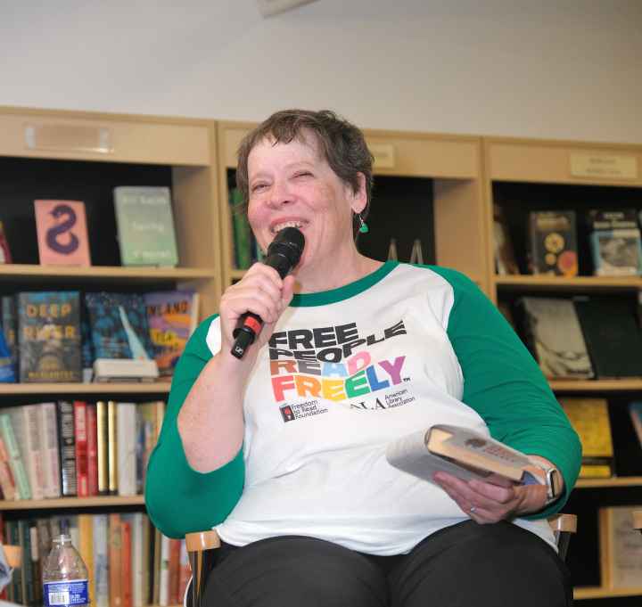 white Woman in a white and green tshirt holding a mircophone and a book addressing a crowd in a bookstore