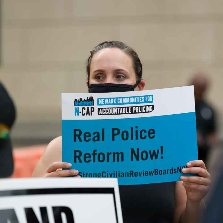 A masked woman holds a sign by Newark Communities for Accountable Policing that reads: Real Police Reform Now!