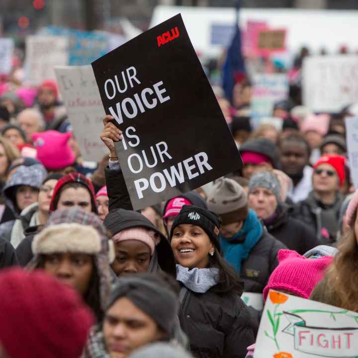 A smiling woman is holding a sign that says "Our voice is our power" in a crowded protest.