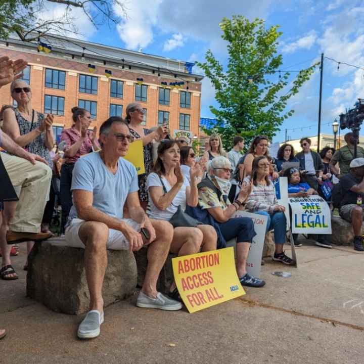 A crowd of people are gathered at a park, clapping and holding signs that read "Abortion Access for All"