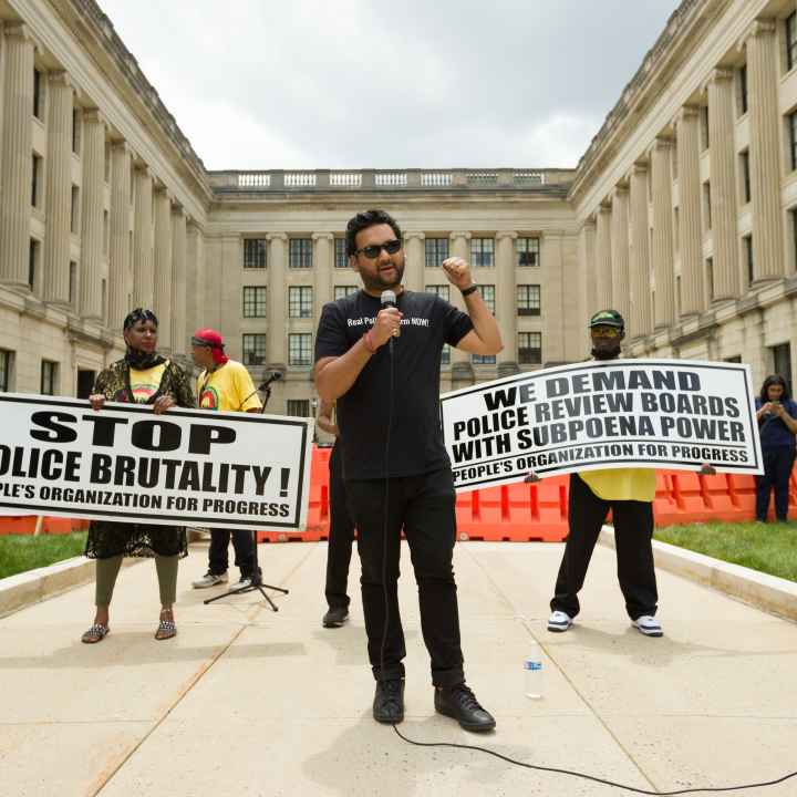 Amol SInha is speaking into a microphone at the State House. Two people are standing behind him holding signs that read "stop police brutality" and "we demand  police review boards with subpoena power"