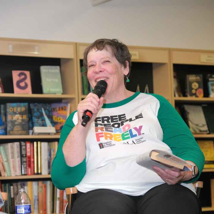white Woman in a white and green tshirt holding a mircophone and a book addressing a crowd in a bookstore