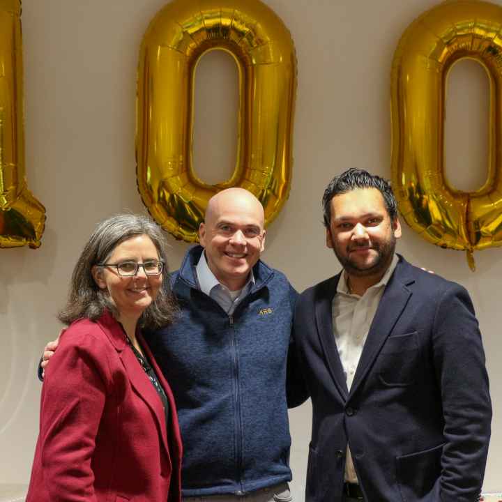 Three people standing in front of ballons of the number 100 in an office conference room