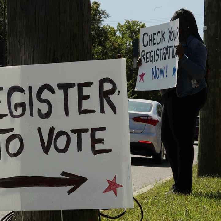 In the foreground, posted on a telephone pole reads a sign "REGISTER TO VOTE", while a woman holds a sign saying "CHECK YOUR REGISTRATION NOW!" is in the background.